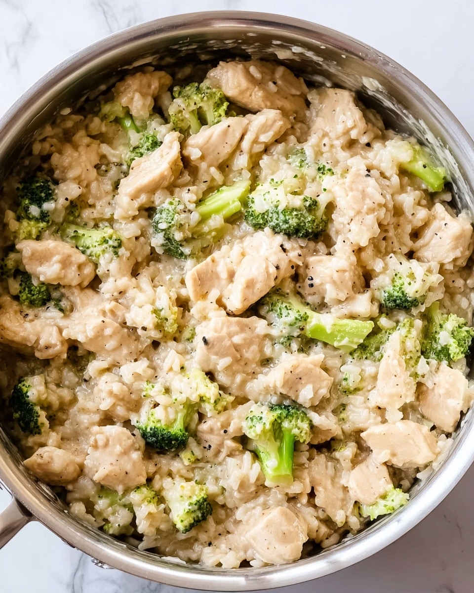 The image shows a close-up of a creamy chicken and broccoli rice dish in a silver pan. The dish has three main layers: tender, light brown pieces of chicken scattered throughout; small, bright green broccoli florets mixed evenly among the chicken; and soft, slightly shiny, light beige rice coating all the ingredients, giving the dish a thick, creamy texture. The mixture looks well blended with some small black pepper specks visible on the chicken and rice. The pan sits on a white marbled surface. photo taken with an iphone --ar 4:5 --v 7