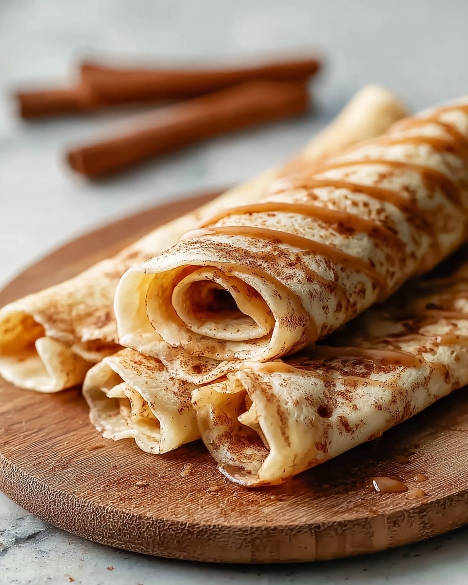 Three rolled crepes are placed side by side on a round white plate, each crepe showing a light golden color with a soft and thin texture. They are generously drizzled with smooth caramel sauce in diagonal lines across the top, and sprinkled with a dusting of fine brown cinnamon powder. Next to the plate, on the white marbled surface, there is a short cinnamon stick and a small white bowl filled with cinnamon powder. A soft pink cloth is partially visible under the plate. The whole scene looks warm and inviting, with the crepes neatly arranged and the toppings adding texture and color contrast. photo taken with an iphone --ar 4:5 --v 7
